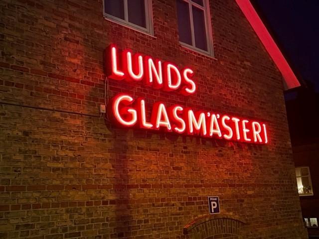 Neonskylt i rött med texten Lunds Glasmästeri mot en gul tegelvägg