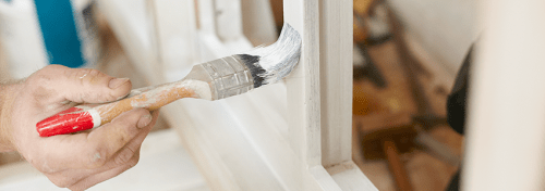En hand håller en pensel som stryker vit färg på en fönsterbåge